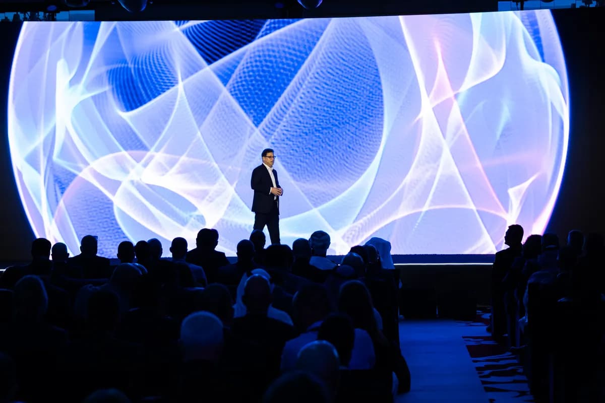 A keynote speaker stands on stage addressing a seated audience during the Deloitte Annual Partners Meeting, silhouetted against a massive LED screen displaying abstract blue and white digital waves.